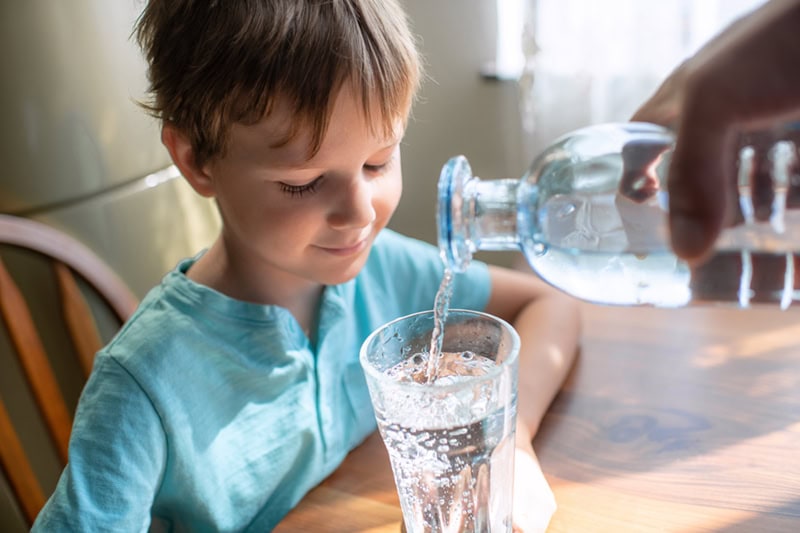 A young boy watching attentively as fresh water is poured into a clear glass sitting on a wooden table.